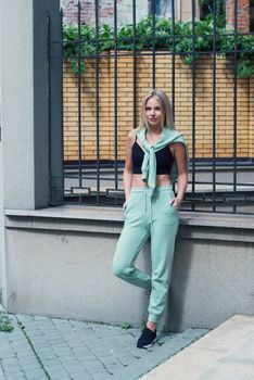 Stylish beautiful young blond woman in a light green tracksuit posing on the street. Attractive girl model posing outdoors. Fitness lady