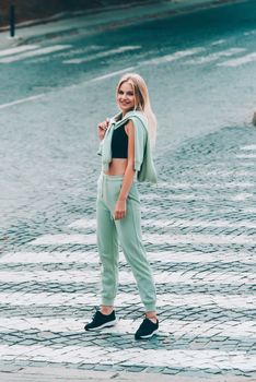 Stylish beautiful young blond woman in a light green tracksuit posing on the street. Attractive girl model posing outdoors. Fitness lady