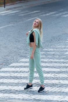 Stylish beautiful young blond woman in a light green tracksuit posing on the street. Attractive girl model posing outdoors. Fitness lady