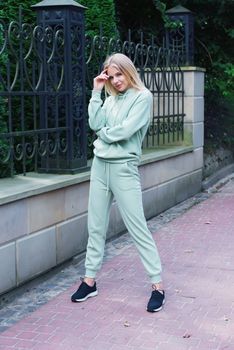 Stylish beautiful young blond woman in a light green tracksuit posing on the street. Attractive girl model posing outdoors. Fitness lady