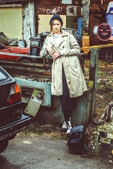 Beautiful young stylish blonde woman wearing long beige coat, white boots, black hat and backpack posing through the city streets. Trendy casual outfit. Street fashion. author's toning