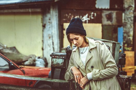 Beautiful young stylish blonde woman wearing long beige coat, white boots, black hat and backpack posing through the city streets. Trendy casual outfit. Street fashion. author's toning