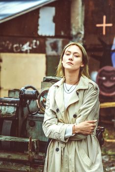 Beautiful young stylish blonde woman wearing long beige coat, white boots, and backpack posing through the city streets. Trendy casual outfit. Street fashion. author's toning