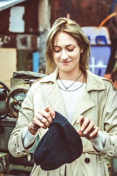 Beautiful young stylish blonde woman wearing long beige coat, white boots, black hat and backpack posing through the city streets. Trendy casual outfit. Street fashion. author's toning