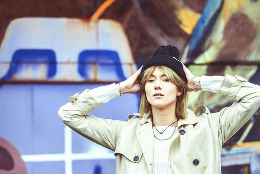 Beautiful young stylish blonde woman wearing long beige coat, white boots, black hat posing through the city streets. Trendy casual outfit. Street fashion. author's toning