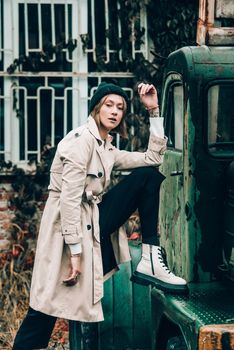 Beautiful young stylish blonde woman wearing long beige coat, white boots and black hat posing on the old green truck. Trendy casual outfit. Selective focus, grain. Street fashion. author's toning