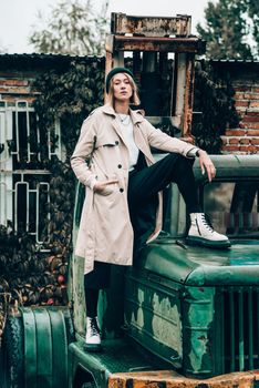 Beautiful young stylish blonde woman wearing long beige coat, white boots and black hat posing on the old green truck. Trendy casual outfit. Selective focus, grain. Street fashion. author's toning