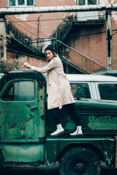 Beautiful young stylish blonde woman wearing long beige coat, white boots and black hat posing on the old green truck. Trendy casual outfit. Selective focus, grain. Street fashion. author's toning