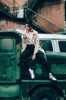 Beautiful young stylish blonde woman wearing long beige coat, white boots and black hat posing on the old green truck. Trendy casual outfit. Selective focus, grain. Street fashion. author's toning