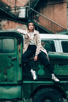 Beautiful young stylish blonde woman wearing long beige coat, white boots and black hat posing on the old green truck. Trendy casual outfit. Selective focus, grain. Street fashion. author's toning