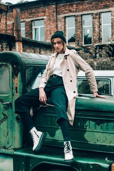 Beautiful young stylish blonde woman wearing long beige coat, white boots and black hat posing on the old green truck. Trendy casual outfit. Selective focus, grain. Street fashion. author's toning