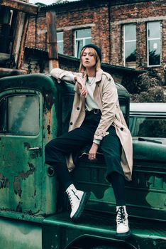 Beautiful young stylish blonde woman wearing long beige coat, white boots and black hat posing on the old green truck. Trendy casual outfit. Selective focus, grain. Street fashion. author's toning