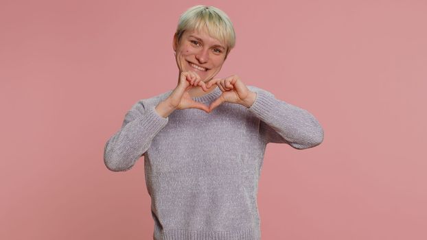 Woman in love. Smiling woman with short blonde hair makes heart gesture demonstrates love sign expresses good feelings and sympathy. Young girl isolated on pink background. Lgbt gay lesbian people
