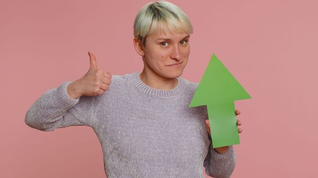 Young woman winner holding arrow sign pointing up, career growth and money exchange increase. Girl showing up indicating successful development progress on pink studio background. Lgbt gay lesbian