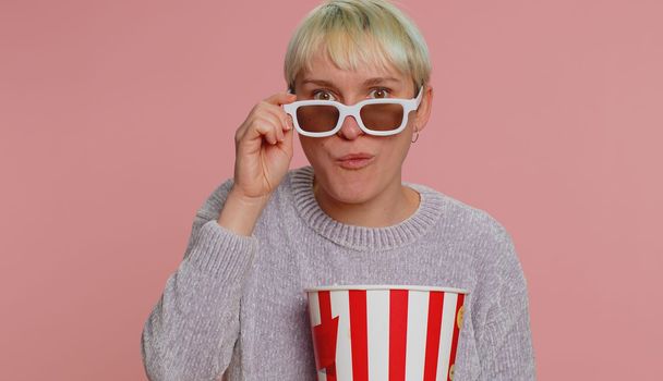 Excited woman in 3D glasses eating popcorn, watching interesting tv serial, sport game, film, online social media movie content. Young girl enjoying domestic entertainment on pink studio background
