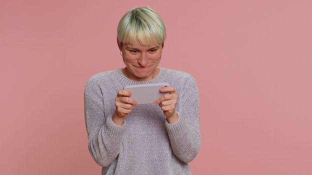 Worried funny addicted woman with short hair enthusiastically playing racing video games on mobile phone. Young girl using smartphone gadget app with drive simulator isolated on pink studio background