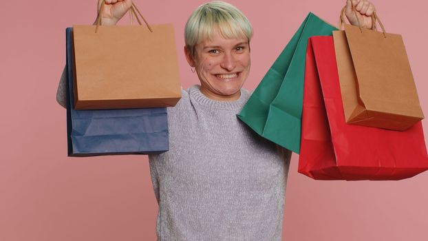 Happy tourist woman with short hair showing shopping bags, advertising discounts, smiling looking amazed with low prices, shopping on Black Friday holidays. Young millennial girl on pink background