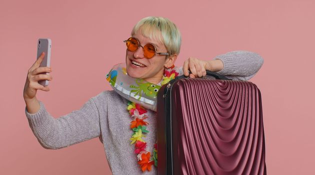 Traveler blogger woman with short hair with swimming ring and luggage, taking selfie on smartphone, make video call online. Young girl on summer holiday vacation, trip. Tourism. Pink studio background