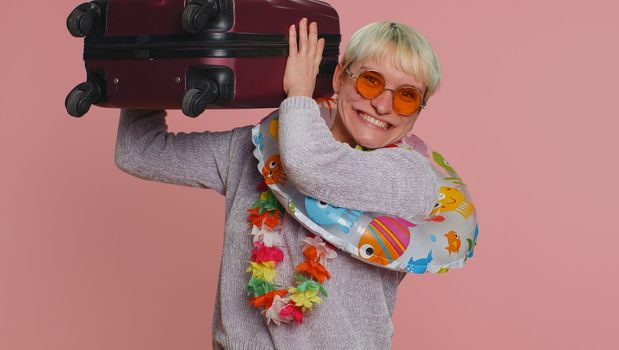 Traveler tourist woman with short hair celebrating, holding luggage, swimming ring. Concept summer vacation, journey, trip to seaside. Air flight journey. Young girl in sunglasses on pink background