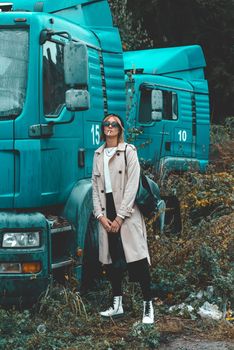 Beautiful young stylish blonde woman wearing long beige coat, white boots and black hat posing smoking a cigarette near the blue truck. Trendy casual outfit. Selective focus, grain. Street fashion.