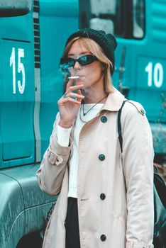 Beautiful young stylish blonde woman wearing long beige coat, white boots and black hat posing smoking a cigarette near the blue truck. Trendy casual outfit. Selective focus, grain. Street fashion. author's toning