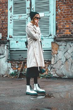 young stylish woman wearing long beige coat, white boots, black hat and backpack posing near blue jalousie door Trendy casual outfit. Selective focus, grain Street fashion. author's toning