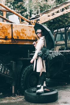 young stylish woman wearing long beige coat, white boots, black hat and umbrella posing near old truck crane. Trendy casual outfit. Selective focus, grain Street fashion. author's toning