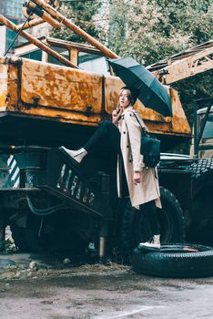 young stylish woman wearing long beige coat, white boots, black hat and umbrella posing near old truck crane. Trendy casual outfit. Selective focus, grain Street fashion. author's toning