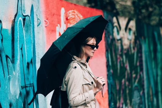 Beautiful young stylish blonde woman wearing long beige coat, white boots, black hat, umbrella and backpack posing through the city streets. Trendy casual outfit. Selective focus, grain