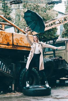 young stylish woman wearing long beige coat, white boots, black hat and umbrella posing near old truck crane. Trendy casual outfit. Selective focus, grain Street fashion. author's toning