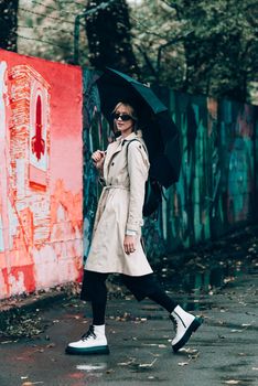 Beautiful young stylish blonde woman wearing long beige coat, white boots, black hat, umbrella and backpack posing through the city streets. Trendy casual outfit. Selective focus, grain