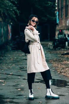 Beautiful young stylish blonde woman wearing long beige coat, white boots, black hat, umbrella and backpack posing through the city streets. Trendy casual outfit. Selective focus, grain