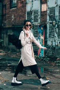 Beautiful young stylish blonde woman wearing long beige coat, white boots, black hat, umbrella and backpack posing through the city streets. Trendy casual outfit. Selective focus, grain