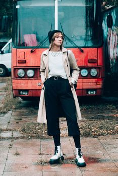 Beautiful young stylish blonde woman wearing long beige coat, white boots and black hat posing on the old red bus. Trendy casual outfit. Selective focus, grain. Street fashion. author's toning