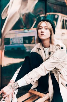 Beautiful young stylish blonde woman wearing long beige coat, white boots, black hat posing through the city streets. Trendy casual outfit. Street fashion. author's toning