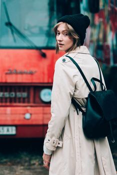 Beautiful young stylish blonde woman wearing long beige coat, white boots and black hat posing on the old red bus. Trendy casual outfit. Selective focus, grain. Street fashion. author's toning