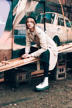Beautiful young stylish blonde woman wearing long beige coat, white boots, black hat posing through the city streets. Trendy casual outfit. Street fashion. author's toning