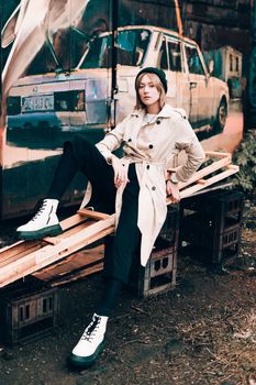 Beautiful young stylish blonde woman wearing long beige coat, white boots, black hat posing through the city streets. Trendy casual outfit. Street fashion. author's toning