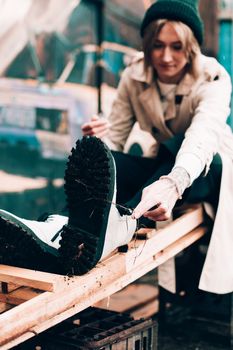 Beautiful young stylish blonde woman wearing long beige coat, white boots, black hat posing through the city streets. Trendy casual outfit. Street fashion. author's toning