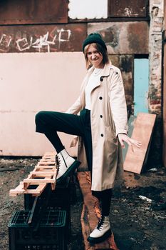 Beautiful young stylish blonde woman wearing long beige coat, white boots, black hat posing through the city streets. Trendy casual outfit. Street fashion. author's toning