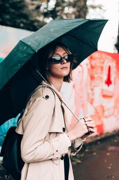 Beautiful young stylish blonde woman wearing long beige coat, white boots, black hat, umbrella and backpack posing through the city streets. Trendy casual outfit. Selective focus, grain