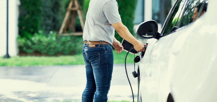 Progressive asian man install cable plug to his electric car with home charging station in the backyard. Concept use of electric vehicles in a progressive lifestyle contributes to clean environment.