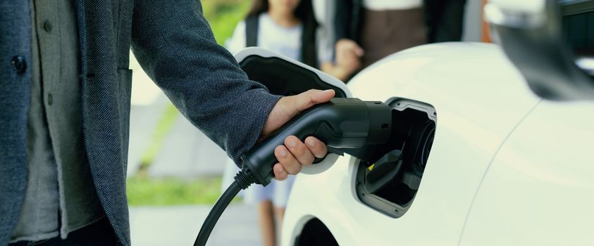 Focus closeup hand charging electric car, insert charger device into electric vehicle as progressive lifestyle concept of alternative green energy technology with blurred family in the background.