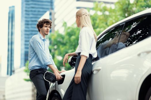 Progressive businessman and businesswoman with electric car connected to charging station before driving around city center. Eco friendly rechargeable car powered by alternative clean energy.