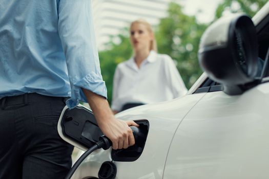 Closeup focus hand insert EV charger to electric vehicle at public charging point in car park with blur business people in backdrop, eco-friendly lifestyle by rechargeable car for progressive concept.