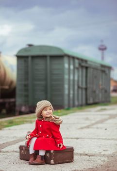 charming baby in a red coat with a suitcase is waiting for a train on the platform