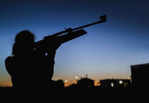 silhouette of a girl who at night on the background of houses aim with a rifle