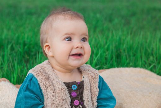 toothless baby in a multi-colored dress sitting on a bedspread on lawn.