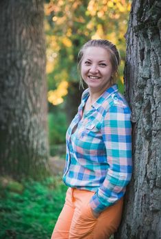 blonde in orange trousers and a plaid shirt smiling in the forest .