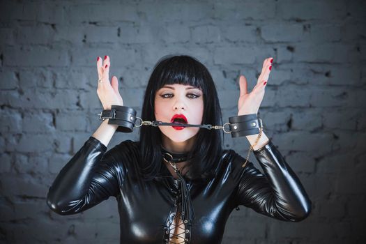 beautiful brunette in a black leather suit and collar gnaws at handcuffs